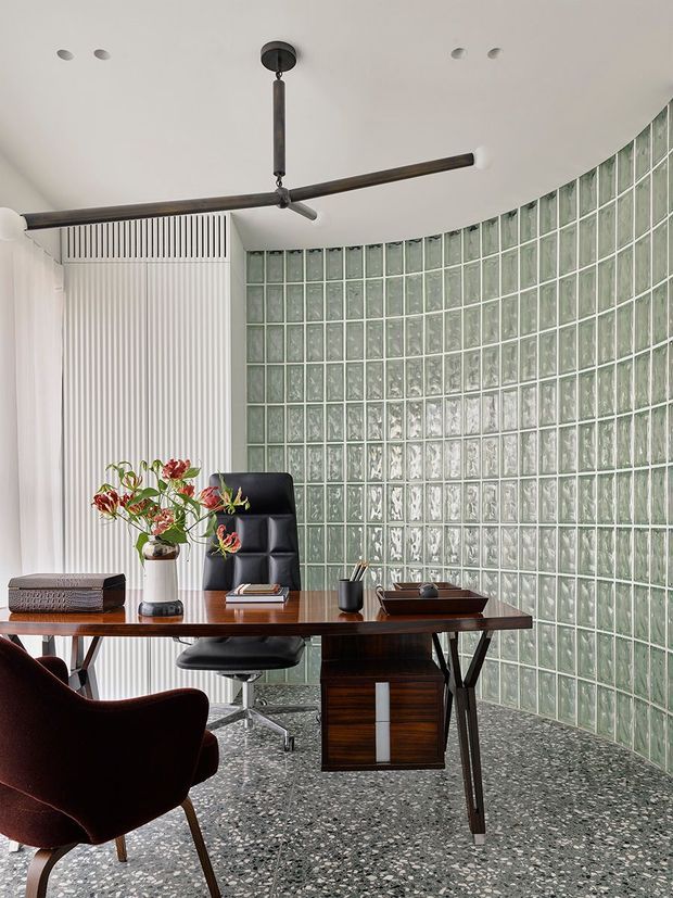A refined home office with a curved wall of translucent green-tinted glass blocks, white fluted wall panelling, a dark timber desk, black leather office chair, burgundy lounge chair, and a sculptural dark ceiling fan. The terrazzo-style floor and restrained palette create a sophisticated mid-century-inspired workspace with abundant diffused light.
