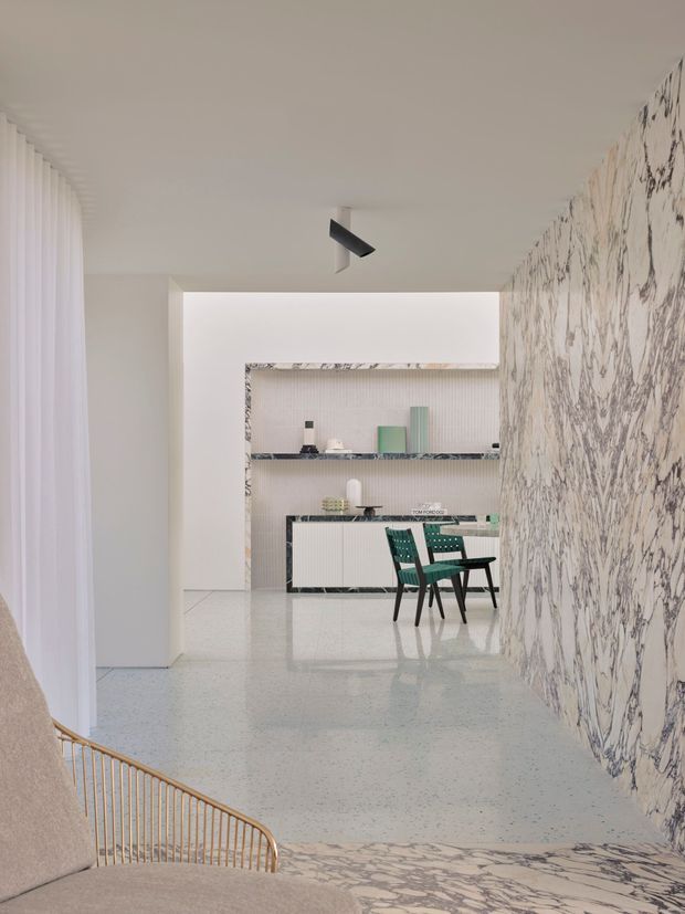 A refined open-plan interior viewed from a lounge area toward a dining and kitchen zone. The space features a pale terrazzo floor, dramatic veined marble wall and stair-side surface, white walls and ceiling, sheer curtains, integrated shelving and cabinetry, and dark green dining chairs around a compact table. A sculptural black ceiling spotlight and warm-toned railing add contrast to the otherwise light, minimalist setting.