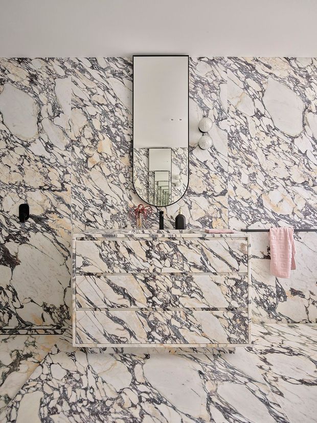 A highly dramatic bathroom clad almost entirely in richly veined white and grey marble, including the walls, floor, vanity front, and benchtop. A floating marble-faced vanity with integrated drawers sits below a tall rounded rectangular mirror, with black tapware and small wall-mounted globe lights. A pink towel provides a soft accent against the monochromatic stone.