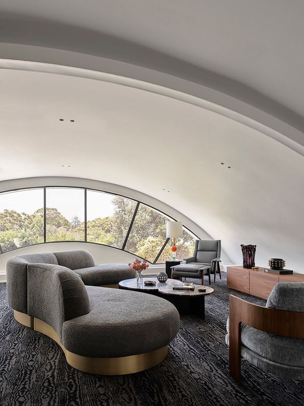 A sophisticated curved living room beneath a sculptural arched ceiling, featuring panoramic arched glazing overlooking trees, a large rounded grey sectional sofa, matching lounge chairs, black round coffee tables, a timber media console, patterned dark carpet, and sculptural decorative objects. The space has a refined contemporary feel with strong mid-century-inspired furniture forms and warm metallic detailing.