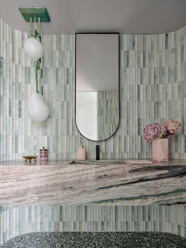 A highly stylised bathroom vanity with vertically patterned pale green and grey wall tiles, a dramatic veined stone benchtop, and a large rounded-rectangle mirror with a black frame. Two sculptural white pendant lights hang from a green ceiling mount, while pink accessories and flowers add colour. A terrazzo-like dark floor is visible at the bottom.