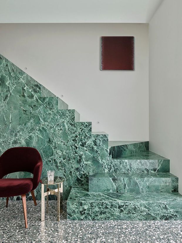 A refined interior stairway clad in dramatic green veined stone, with matching stone risers, treads, and side wall. The staircase rises beside a light neutral plaster wall with small circular details and a dark red framed artwork. A burgundy upholstered chair and small glass-topped side table sit on a grey terrazzo floor, creating a bold, sophisticated composition.