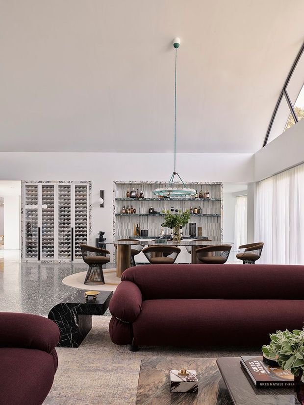 A refined open-plan living and dining interior with a dramatic burgundy curved sofa in the foreground, a matching lounge chair, a dark marble coffee table, and a dining table with sculptural wooden chairs. Behind the dining area is a large built-in bar or display wall with open shelving and a textured backsplash, while extensive sheer glazing runs along the right side. A large suspended sculptural pendant light hangs over the dining table. The room has polished speckled flooring, pale walls, high ceilings, and a sophisticated contemporary aesthetic.