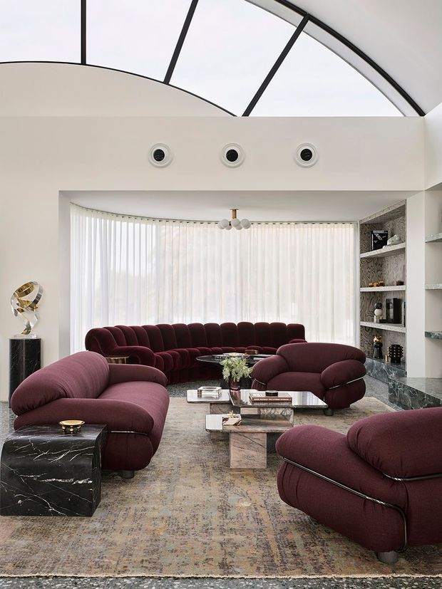 A luxurious contemporary living room with a dramatic curved glazed ceiling, expansive sheer-covered windows, deep burgundy upholstered curved sofas and armchairs, marble side tables, a patterned area rug, and built-in open shelving. The room has a bright, refined atmosphere with strong sculptural furniture forms and a high curved ceiling.