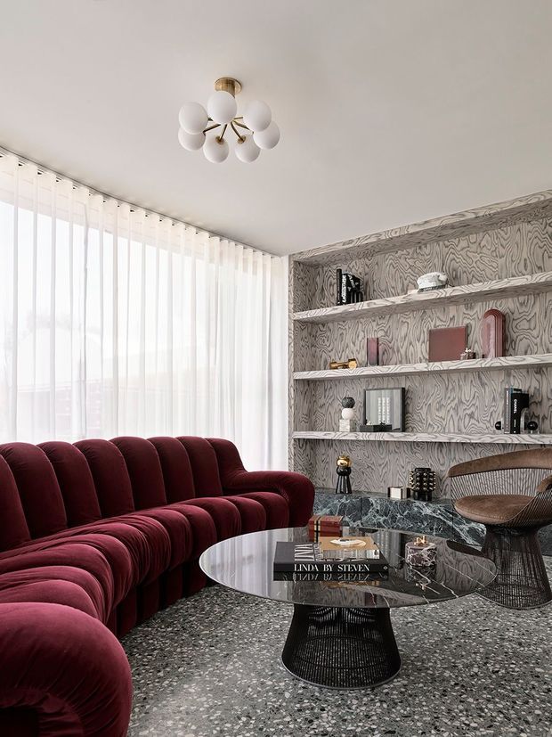 A sophisticated living room featuring a deep burgundy scalloped sectional sofa, round glass coffee table with a dark sculptural base, terrazzo flooring, and a marble-clad low console. The walls are covered in a dramatic grey patterned finish with integrated open shelving displaying books and decorative objects. Sheer floor-to-ceiling curtains provide abundant filtered daylight, while a sculptural white globe chandelier adds a playful focal point. A dark woven lounge chair completes the room.