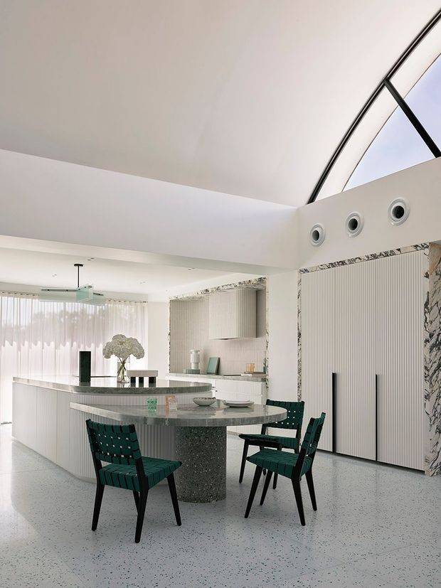 Bright, high-end contemporary kitchen and dining area with a large curved stone island, integrated white cabinetry, green upholstered dining chairs, terrazzo flooring, fluted wall panelling, marble accents, and a dramatic glazed roof opening with exposed dark framing. Circular ceiling vents and abundant natural light add to the refined architectural character.
