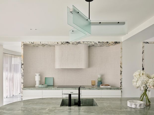 Refined contemporary kitchen viewed from behind a large stone island with an integrated sink and black tapware. The room features pale fluted cabinetry and wall panelling, a matching fluted rangehood, dramatic veined stone trim around the rear cabinetry, and a sculptural glass pendant light. Soft neutral finishes and carefully styled accessories create a luxurious, cohesive interior.