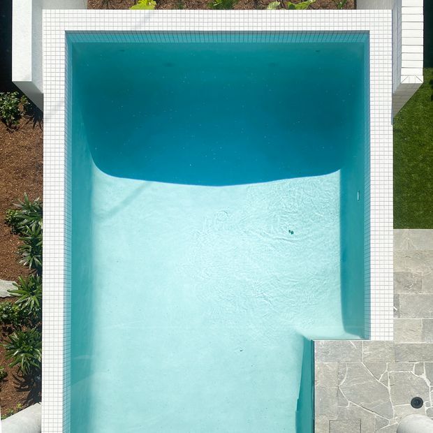 Top-down aerial view of a rectangular swimming pool with a curved transition between deep blue and shallow pale aqua water. The pool is bordered by a white small-format tiled coping, with pale stone paving and landscaped planting around the edges.