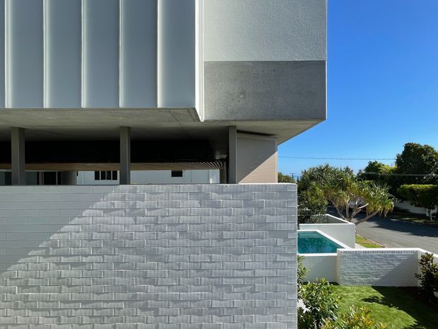 Exterior view of a contemporary multi-level residence with strong horizontal and cantilevered forms, pale textured cladding, exposed concrete soffits, and slender dark-framed openings. A light brick privacy wall encloses a landscaped yard and compact swimming pool, with neighboring suburban context visible beyond.