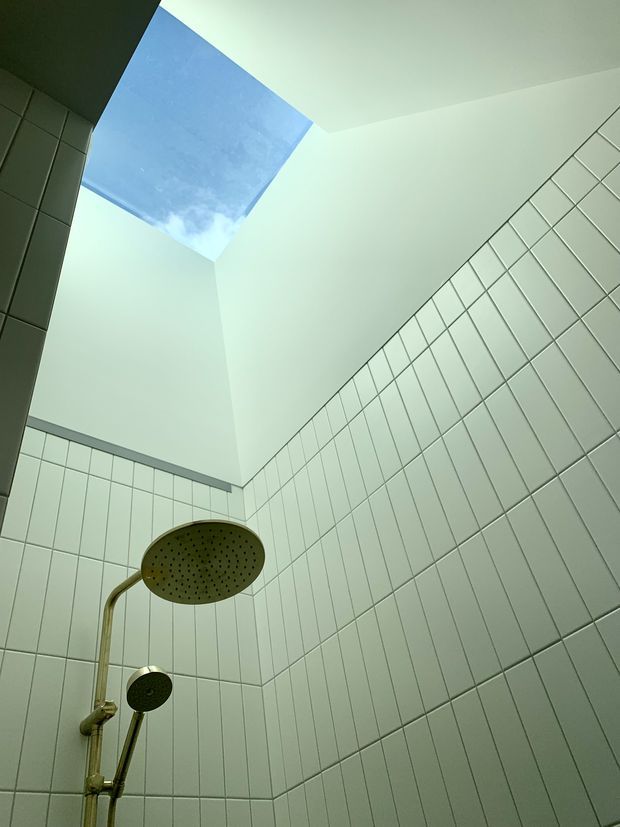 Upward-looking view of a tiled shower enclosure with pale rectangular wall tiles, a large rainfall showerhead, handheld shower, and brass-toned plumbing. A skylight above the shower brings in natural light and reveals blue sky.