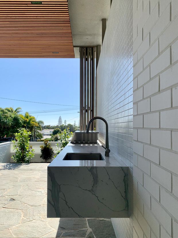 A contemporary outdoor kitchen or entertaining area beside a white tiled masonry wall. The scene features a built-in stone benchtop with an integrated black sink and curved tap, timber slatted overhead lining, dark vertical screening, and a paved terrace overlooking landscaped residential surroundings.
