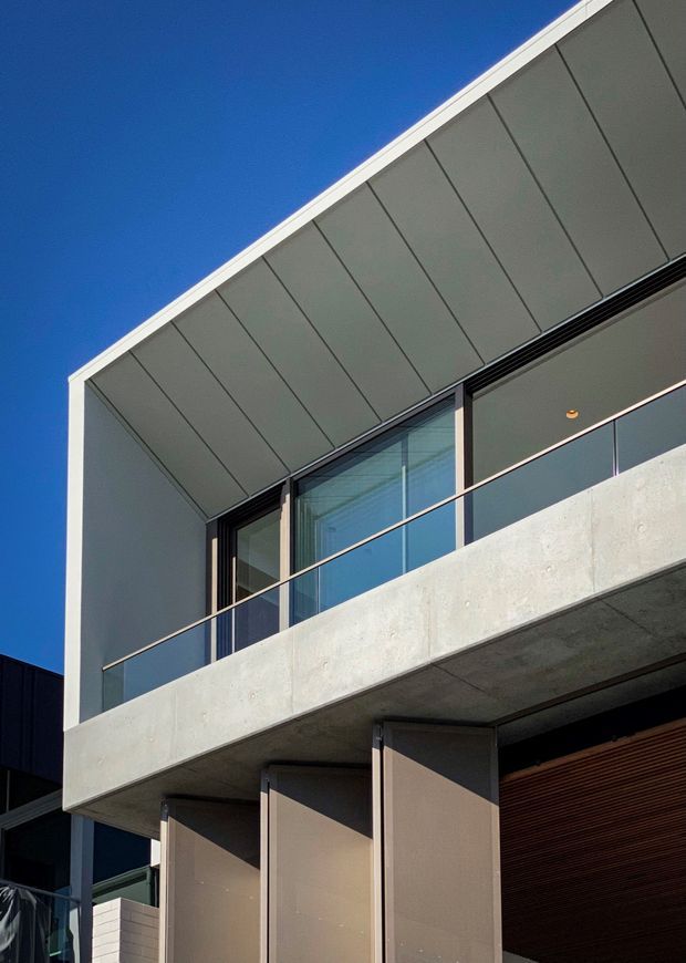 Close-up exterior architectural view of a contemporary multi-level building with a strong cantilevered concrete balcony, full-height glazing, glass balustrade, dark metal framing, and a large roof overhang lined with segmented panels. The facade combines pale exposed concrete, dark cladding, and warm timber-look screening beneath a vivid blue sky.