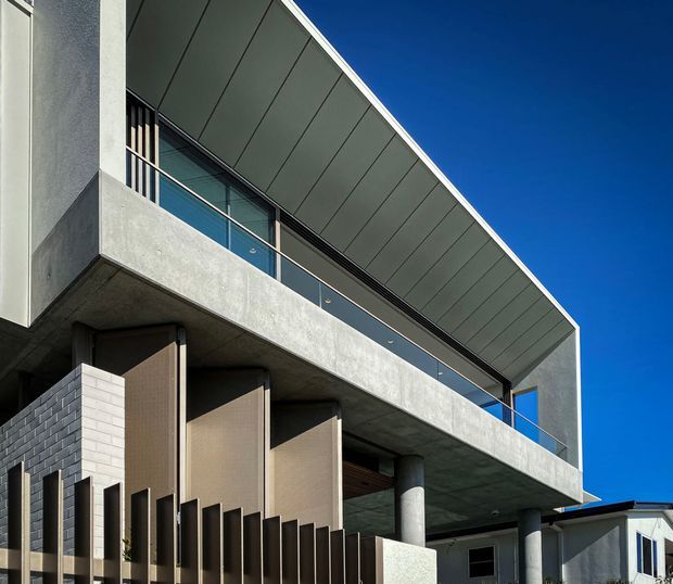 Contemporary multi-level residential facade photographed from below, featuring a projecting concrete balcony, dark metal soffit and roofline, glass balustrade, extensive glazing, vertical privacy screens, cylindrical concrete columns, and pale masonry walls against a clear blue sky.