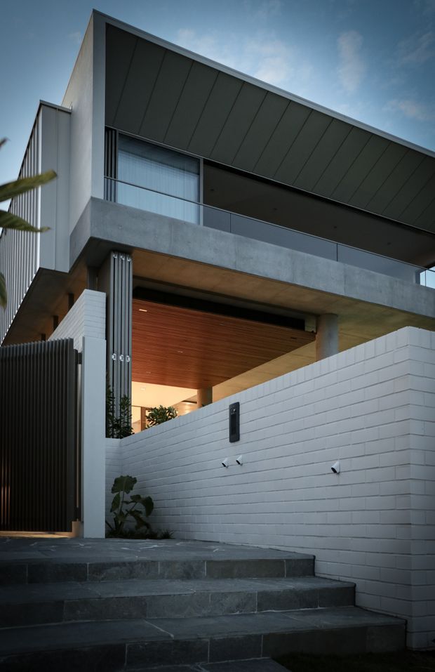 Street-level exterior view of a contemporary multi-storey residence. The facade combines dark vertical metal screening, pale rendered and painted brick walls, exposed concrete balconies and columns, large glazing, and a broad dark metal skillion-style roof form. A warm timber-lined soffit marks the recessed entry and covered carport or garage approach, reached by dark stone-paved steps. The image has a dramatic, sophisticated dusk atmosphere.