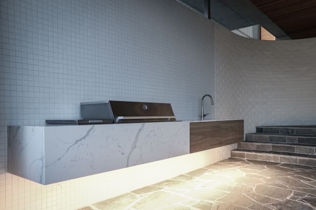 A refined outdoor kitchen with a floating white marble-look island and integrated cabinetry, built-in grill, sink and dark timber panel. Small white square wall tiles wrap the curved wall, while dark stone steps and paving lead upward beside the kitchen. The space has a contemporary, minimalist architectural character with concealed under-cabinet lighting and a partially covered ceiling.