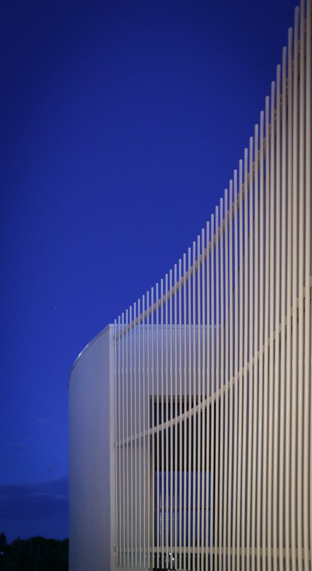 A dramatic dusk exterior view of a contemporary building, dominated by a sweeping screen of closely spaced vertical white fins. The fins rise in a curved, ascending profile and partially veil a white multi-level facade with dark glazing or openings behind them. A deep cobalt-blue evening sky provides strong contrast, emphasizing the building's sculptural geometric form.