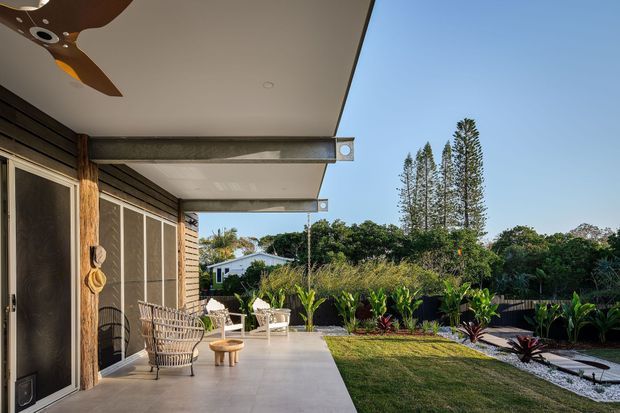 A covered outdoor terrace beside a timber-clad home, with a broad concrete patio, ceiling-mounted timber-blade fan, screened sliding doors, woven lounge chairs, and a small timber stool. The terrace opens onto a landscaped garden with lawn, tropical planting, a gravel path, and tall ornamental trees under a clear blue sky. The setting has a relaxed, warm-climate indoor-outdoor character.