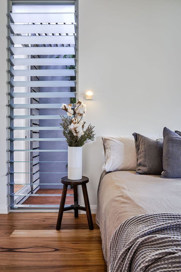 A calm, contemporary bedroom corner with a neatly made bed, layered neutral and grey pillows, and a textured grey throw. Warm timber flooring contrasts with pale walls and a tall aluminium louvred window or shutter system. A small dark timber stool supports a white vase of dried branches, while a compact wall light adds a warm glow.