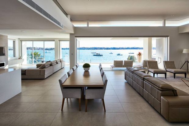 A spacious open-plan waterfront living and dining area with floor-to-ceiling glazing and broad views across the water. The room features large tiled flooring, neutral walls, a long timber dining table with upholstered chairs, and multiple brown leather sofas and armchairs. The bright, minimal interior opens directly toward the scenic waterfront.