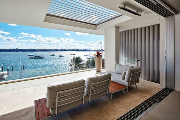 A bright waterfront outdoor living area with a built-in louvred roof and retractable glazed enclosure. It contains a lounge seating arrangement facing the water, tiled flooring, vertical privacy blinds, and a timber-topped coffee table. Boats and a distant shoreline are visible beyond the balustrade.