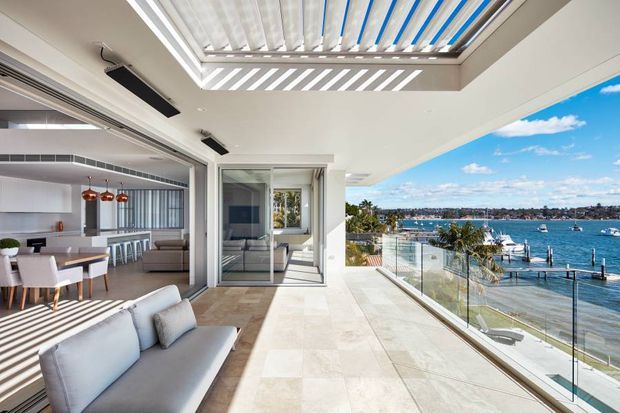 A bright, contemporary waterfront apartment terrace opening directly into an open-plan living and dining area with a sleek kitchen. The large outdoor entertaining space has pale stone-look paving, frameless glass balustrades, and a retractable louvred roof providing striped sunlight. Inside are neutral upholstered furnishings, a dining table, island seating, and pendant lights. The terrace overlooks a marina or calm coastal inlet with a private dock and boats.