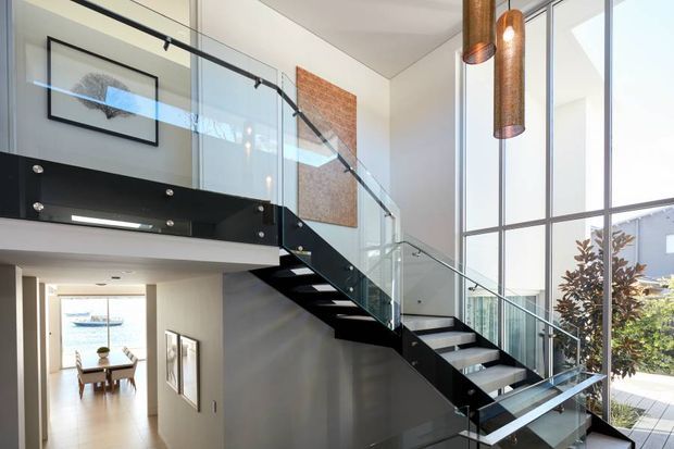 Bright contemporary interior featuring a dramatic open-riser staircase with black steel stringers and balustrade, clear glass guarding, and timber treads. The stair rises beside expansive floor-to-ceiling glazing, with a mezzanine or upper landing, pendant lights, and a dining area visible beyond.