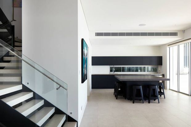 Bright contemporary open-plan dining and living area with a dramatic floating staircase at left, glass balustrade, pale tiled flooring, and a long dark dining table with stools. A minimalist black built-in feature wall and reflective inset sit at the rear, while full-height glazed doors bring in abundant daylight.
