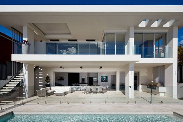 Modern multi-level residence viewed from the pool, featuring a crisp white rendered facade, extensive floor-to-ceiling glazing, a projecting upper balcony, open-plan living and dining areas, and a broad outdoor entertaining terrace. The architecture includes strong horizontal lines, glass balustrades, a flat roof, exposed white structural columns, and a side stairway.