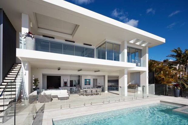 A bright contemporary multi-level residence with strong horizontal lines, white rendered walls, extensive floor-to-ceiling glazing, glass balustrades, and an open-plan ground-floor entertaining area. The home overlooks a turquoise swimming pool and includes an exterior stair, covered terrace, and lush tropical planting at the edges of the frame.