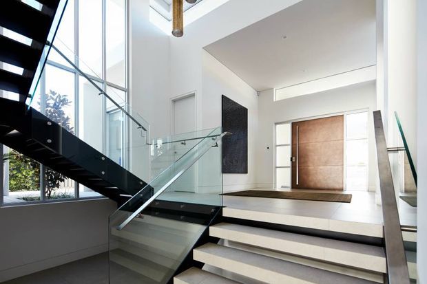 Bright contemporary entry hall with a dramatic split-level staircase. The space features broad white steps, dark timber or black stair structure, clear glass balustrades, large floor-to-ceiling glazing, white walls, and a substantial timber pivot entry door. A dark textured feature wall and exposed timber ceiling detail add contrast.