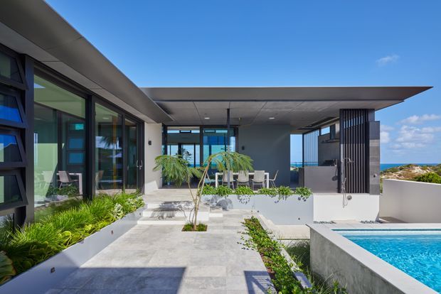 A contemporary flat-roofed residence with an open courtyard terrace and swimming pool. Large dark-framed glazed walls surround the outdoor living area, while a broad cantilevered roof, pale rendered walls, concrete paving, vertical privacy screens and integrated planting create a clean, resort-like composition. The ocean and rocky coastline are visible beyond the property.