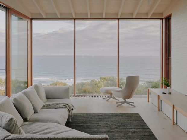 A serene modern living room overlooking the ocean through expansive floor-to-ceiling glazing. The room features a large pale upholstered sectional sofa, a sculptural lounge chair with ottoman, a simple timber media console, a dark textured rug, white-painted brick walls, and a white exposed-beam ceiling. Warm timber window frames and coastal vegetation are visible outside.