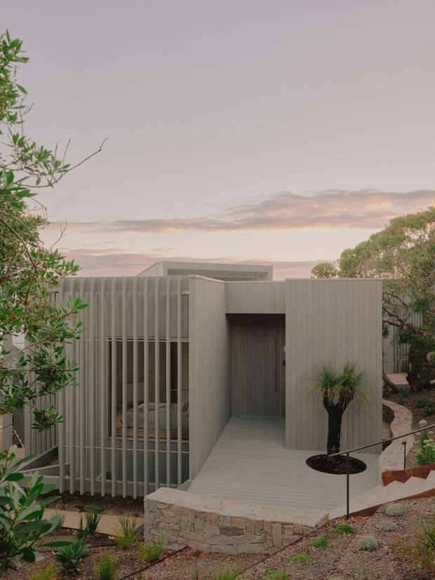 A compact contemporary single-storey dwelling set within landscaped grounds, photographed at dusk. The building has a sculptural flat-roofed form, pale grey vertically ribbed cladding, screened glazing, and a recessed entry reached by a light timber walkway. Stone retaining walls, gravel planting beds, native-style vegetation, and a simple dark handrail define the approach.