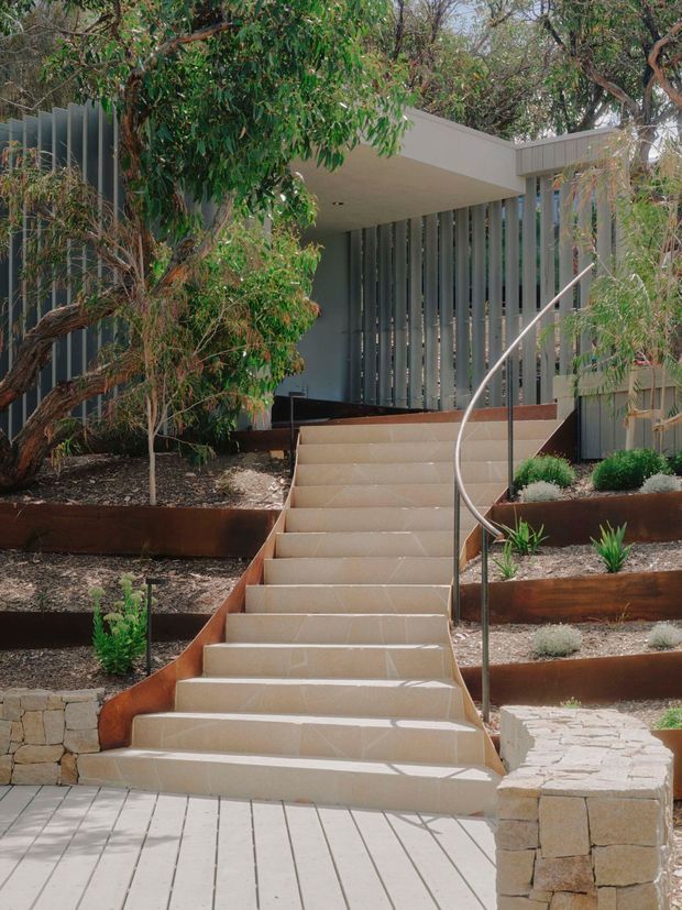 Exterior view of a modern home entrance approached by a broad flight of pale tiled steps. The facade combines dark grey vertical screening and a flat projecting roof canopy, partially concealed by established native-style planting. Rusted steel retaining beds, gravel garden areas, stone retaining walls and a curved metal handrail create a contemporary landscaped approach.