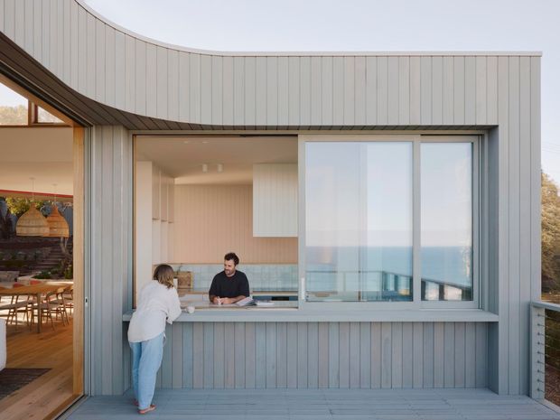 Contemporary coastal home exterior with a curved, vertically clad facade and large glazed opening forming a kitchen servery or counter to an outdoor deck. Through the opening, a light-toned kitchen and dining area are visible, with the ocean horizon beyond. The image also shows timber decking, slim metal-framed glazing, and a relaxed indoor-outdoor connection.