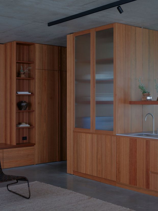 A refined, compact kitchen and integrated storage wall clad in warm vertical timber joinery. The cabinetry includes open display shelving, tall ribbed-glass doors, concealed storage, and a stone benchtop with a curved metal tap. A dark concrete ceiling with a linear track light and polished grey floor create a restrained, contemporary interior; part of a black lounge chair and a neutral rug are visible in the foreground.