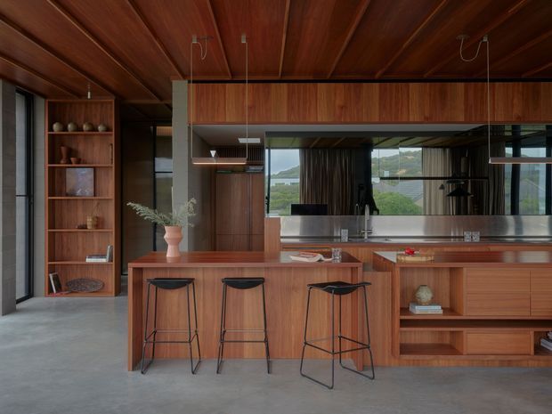 A refined open-plan kitchen dominated by warm timber cabinetry and a timber-lined ceiling. The kitchen features a long island with three black bar stools, integrated cabinetry, open shelving, a dark benchtop, pendant lighting, and a large glazed opening with views beyond. Concrete flooring and dark-framed glazing create a modern, carefully detailed interior.