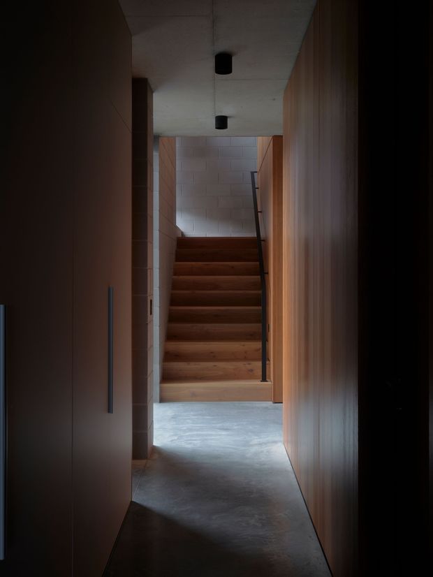 A narrow, moody interior hallway leading to a straight timber staircase. Dark built-in cabinetry or wall panelling lines both sides, while the stair enclosure combines warm timber treads and pale masonry block walls. A smooth raw-looking concrete floor and ceiling, minimal black cylindrical downlights, and a slim dark handrail create a restrained contemporary-industrial atmosphere.