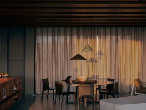 Moody contemporary dining area with a round dining table and dark chairs positioned in front of full-height sheer curtains. The room features a dark timber slatted ceiling, polished concrete or dark flooring, built-in or freestanding timber storage along the left wall, sculptural pendant lighting, and a partial upholstered seating element in the foreground.