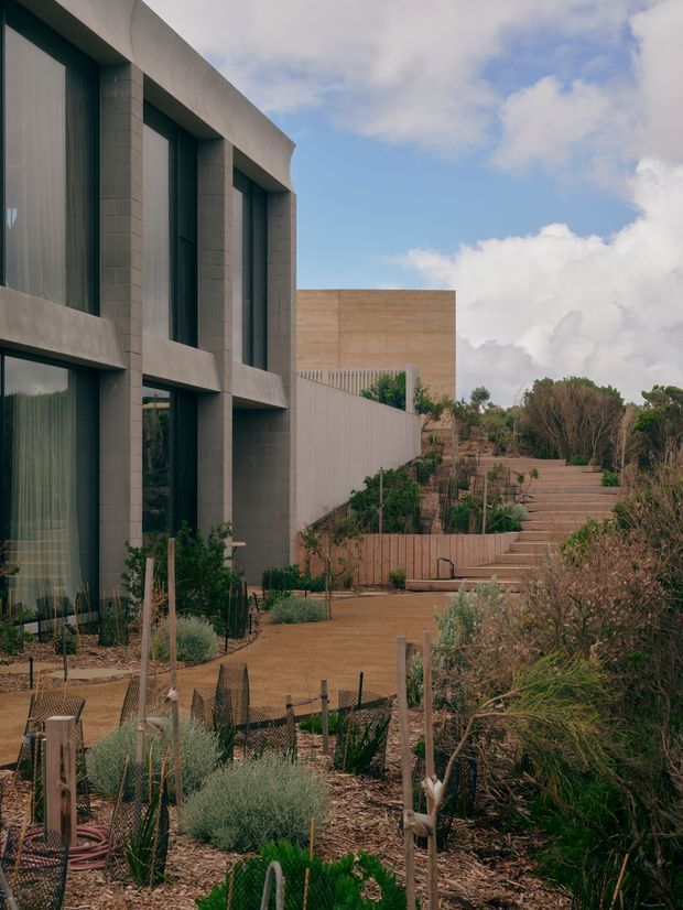 Exterior view of a contemporary multi-storey building beside a landscaped garden. The facade features tall dark-framed glazing set within deep rectangular concrete or masonry piers, with a restrained grey and beige palette. A stepped garden path rises alongside the building, surrounded by native-looking drought-tolerant planting and timber garden structures under a partly cloudy sky.