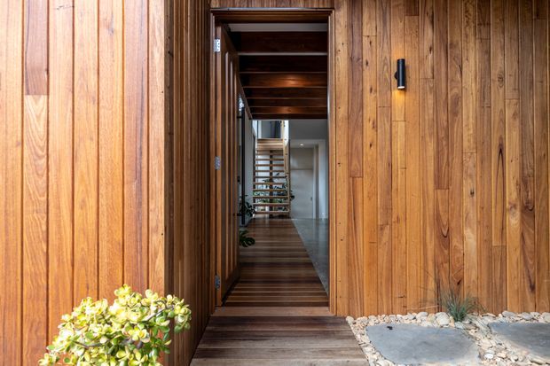 A warm timber-clad residential entryway viewed from outside, with a dark timber pivot or hinged door opening onto a timber-lined passage. The interior beyond includes a timber staircase or screened landing and glimpses of white cabinetry and greenery. Stone pavers, pale gravel, and a small planted shrub frame the threshold, while a slim black wall light adds contrast.