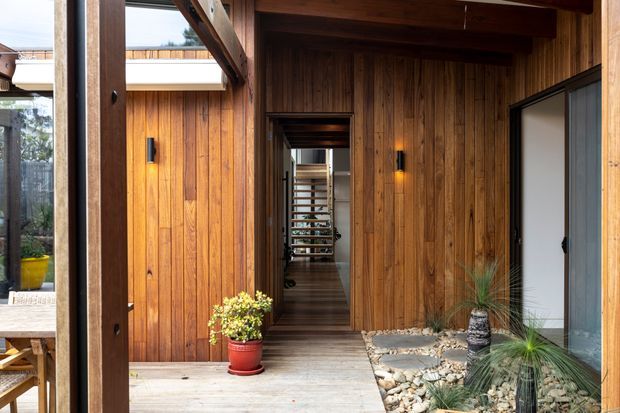 A warm timber-lined entryway or covered courtyard passage with exposed structural beams, dark timber decking, integrated wall lighting, and a central corridor leading deeper into the home. A small rock garden with ornamental grasses borders the right side, while glazing and an outdoor seating area are visible to the left.