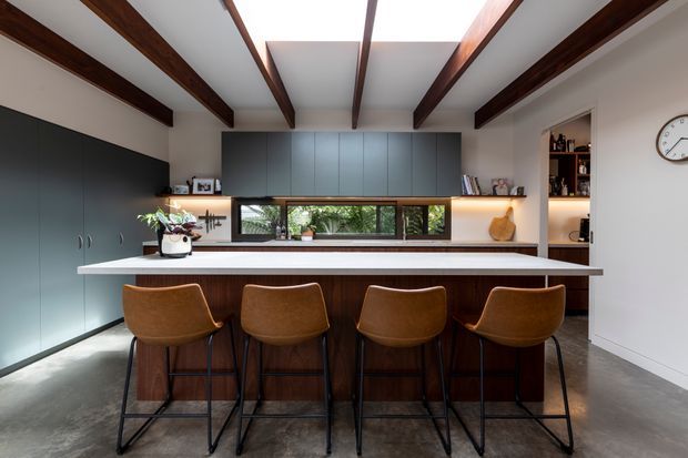 A contemporary kitchen with a long island and four upholstered bar stools. Dark blue-grey full-height cabinetry lines the left wall, while a white island benchtop contrasts with dark timber island cabinetry. The room features exposed dark timber ceiling beams, a horizontal window above the rear benchtop, open shelving, and a neutral grey polished concrete floor.