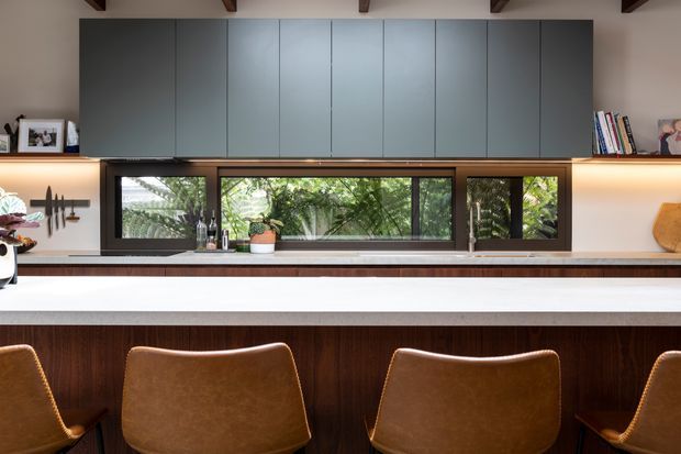 A contemporary kitchen viewed across a large island, with dark timber cabinetry and panelled joinery, a long horizontal window overlooking greenery, warm under-cabinet lighting, exposed timber ceiling beams, and leather counter stools. The palette combines charcoal grey, rich brown timber, white surfaces, and tan leather.