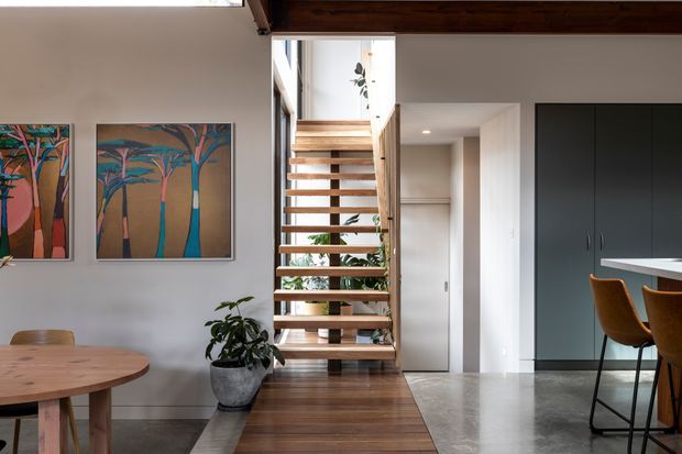 Contemporary open-plan interior featuring a timber floating staircase with open risers, a timber ceiling beam, polished concrete flooring, dark built-in cabinetry, and a partially visible dining area. The staircase is framed by glazing and indoor planting, with colourful tree artwork on the wall and a kitchen or bar area visible to the right.