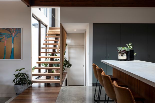 Contemporary open-plan kitchen and entry area with a floating timber staircase, dark full-height kitchen cabinetry, a large light benchtop and timber island base, and a row of brown upholstered bar stools. The space features timber flooring, polished concrete-look flooring around the kitchen, white walls, large glazing, indoor plants, and a dark exposed timber ceiling beam. A hallway and door are visible beyond the stair.