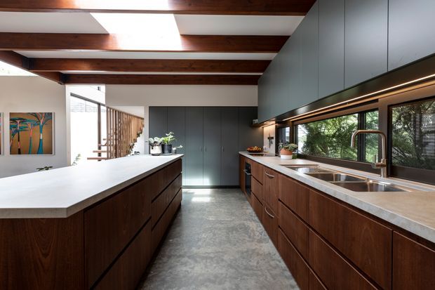 A spacious contemporary galley kitchen with a large central island, dark timber cabinetry, pale stone benchtops, integrated dark grey overhead cabinetry, stainless steel double sink and sculptural tap. Exposed dark timber ceiling beams and clerestory skylights bring warmth and natural light, while full-height windows overlook greenery. The kitchen has a refined mid-century-influenced material palette and generous circulation space.