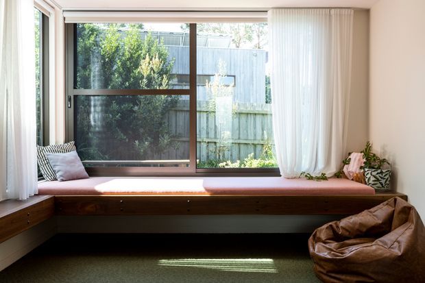 Bright, calm window-seat nook with a built-in timber bench spanning beneath a large framed window. Sheer white curtains, cushions, indoor plants, and a brown leather beanbag create a relaxed residential reading or lounging area. The window overlooks a leafy fenced garden.