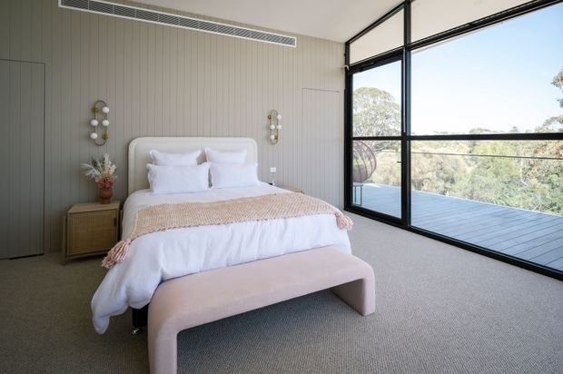 A bright contemporary bedroom with a neatly made upholstered bed, neutral bedding, a soft pink-beige bench, and a woven throw. The room has vertically panelled grey-beige walls, integrated cabinetry, bedside storage, decorative wall sconces, and expansive black-framed glazing opening to a deck and treed landscape.