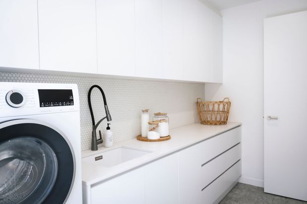 Bright, compact contemporary laundry room with white built-in cabinetry, a front-loading washing machine, integrated sink with black gooseneck tap, pale textured tiled splashback, and a grey floor. A white door opens to the right.